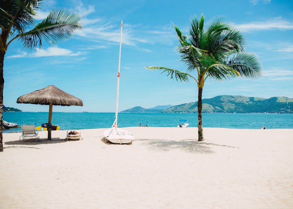 White sand beach with a small sailboat, palm tree, thatched umbrella, lounge chair, and surfboard beside calm turquoise water and green coastal hills